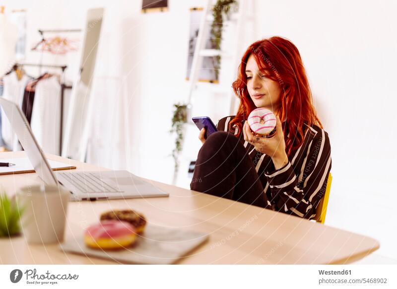 Young businesswoman holding doughnut while using mobile phone sitting at office color image colour image indoors indoor shot indoor shots interior interior view