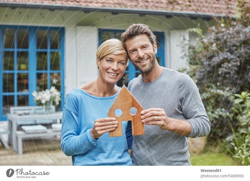 Portrait of smiling couple standing in front of their home holding house model models portrait portraits houses twosomes partnership couples smile building