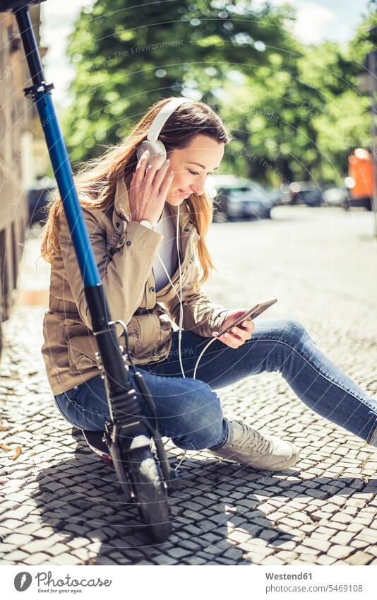 Smiling woman sitting on E-Scooter listening music with headphones and smartphone headset telecommunication telephone telephones cell phone cell phones