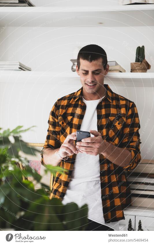 Young man standing at a shelf using cell phone men males Shelve rack racks shelves mobile phone mobiles mobile phones Cellphone cell phones Adults grown-ups