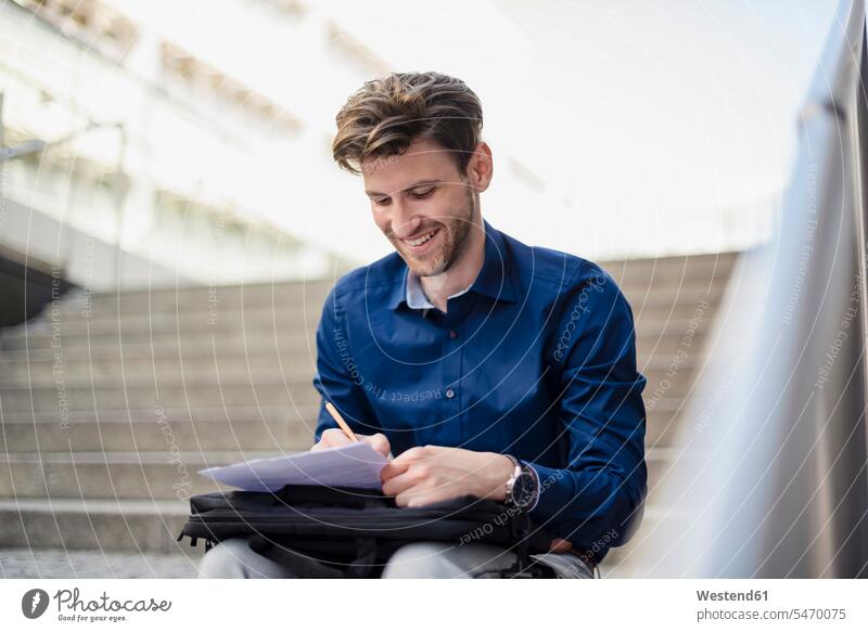 Smiling businessman sitting on stairs in the city taking notes Businessman Business man Businessmen Business men town cities towns making a note note taking