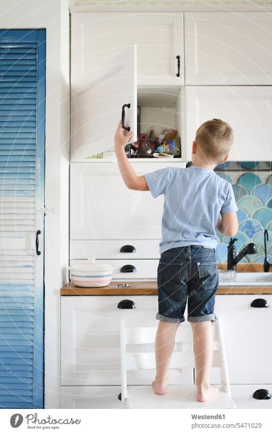 Back view of little boy standing on chair in the kitchen looking into cupboard T- Shirt t-shirts tee-shirt chairs colour colours at home domestic kitchen