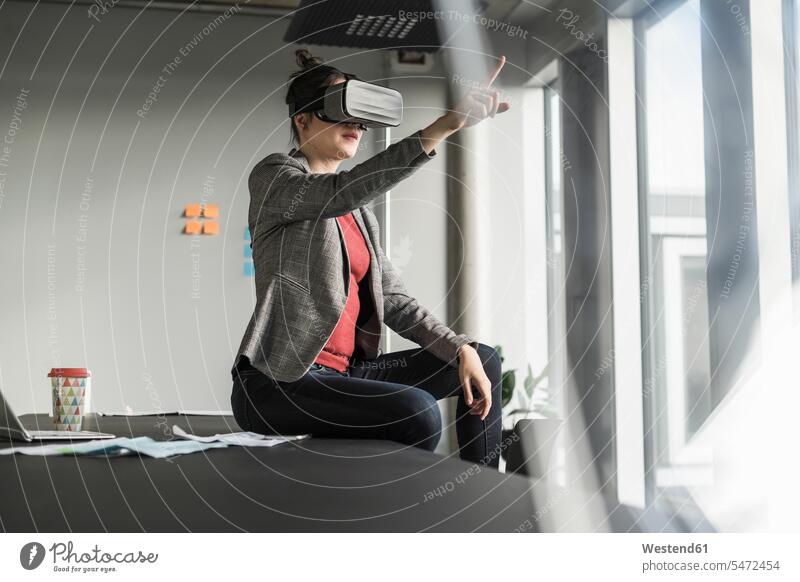 Businesswoman sitting on desk in office wearing VR glasses offices office room office rooms businesswoman businesswomen business woman business women virtual