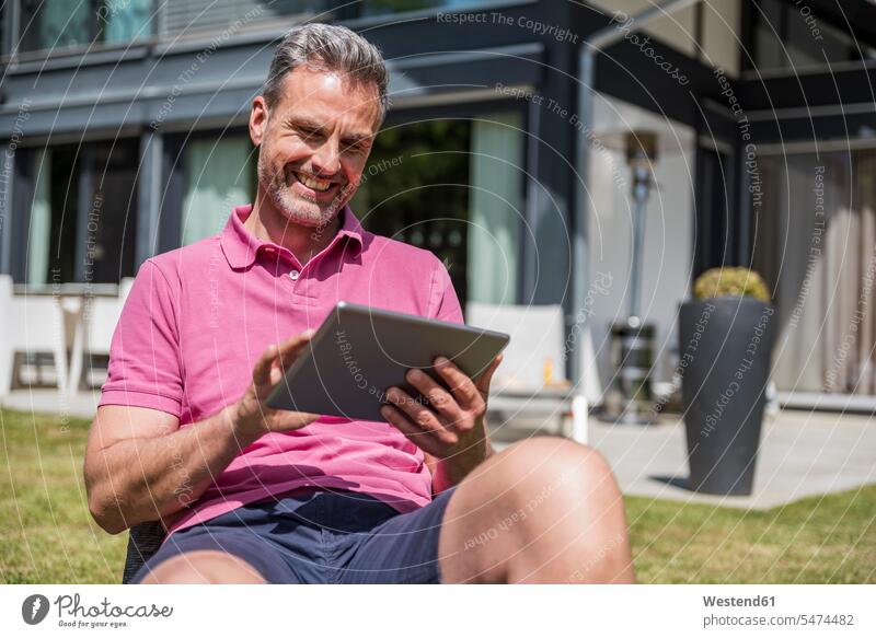 Smiling mature man sitting in garden of his home using a tablet men males smiling smile Seated house houses gardens domestic garden digitizer Tablet Computer