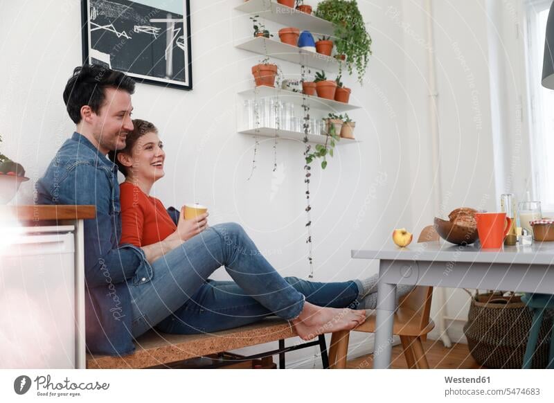 Happy couple sitting in kitchen, leaning against each other twosomes partnership couples happiness happy domestic kitchen kitchens Seated home at home together