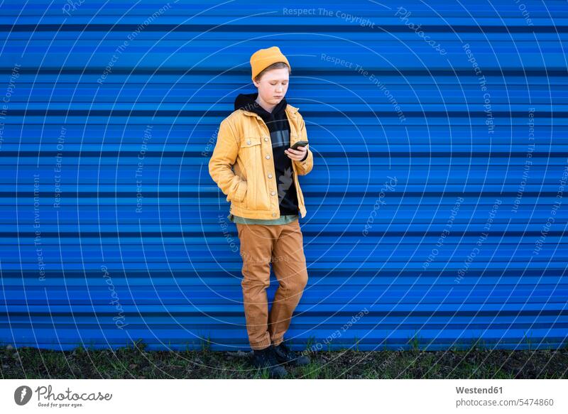 Boy using smart phone while standing against blue shutter color image colour image outdoors location shots outdoor shot outdoor shots day daylight shot