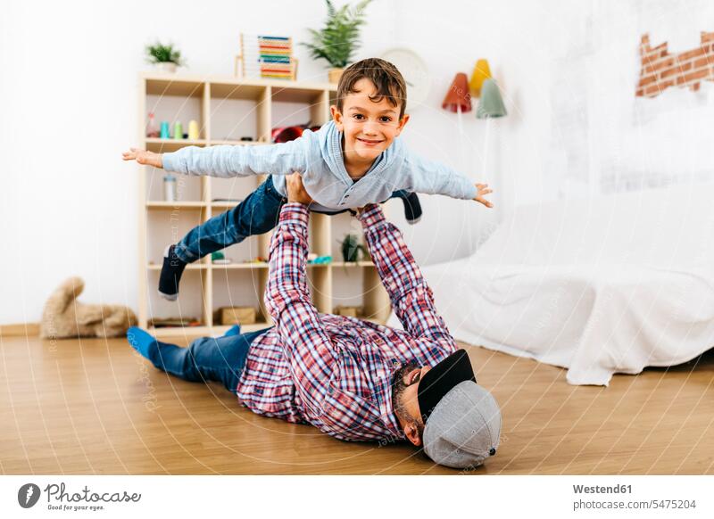 Father lying on floor, holding son aloft, pretending to fly couches settee settees sofa sofas rack racks Shelve shelves smile play delight enjoyment Pleasant