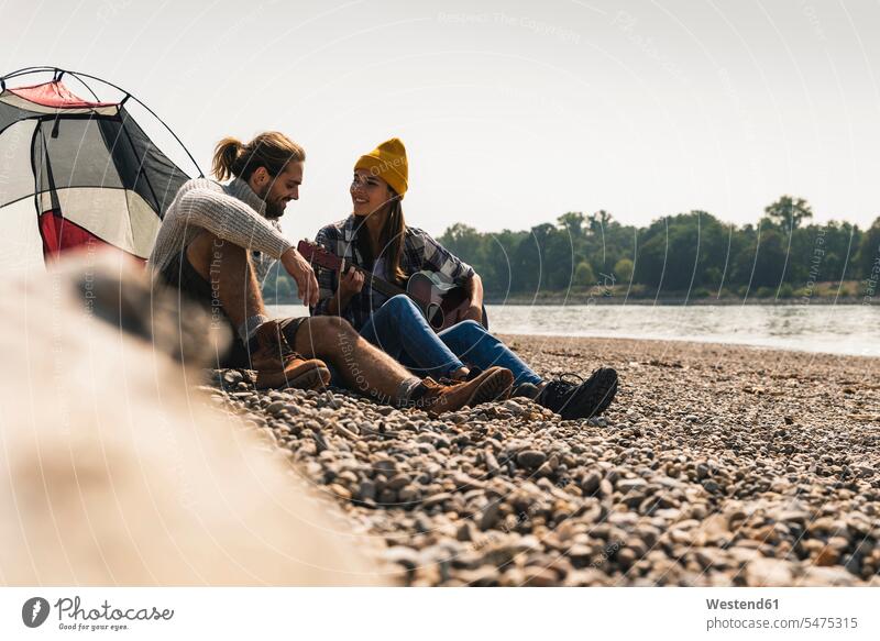 Happy young couple with guitar sitting at a tent at the riverside guitars tents happiness happy riverbank twosomes partnership couples Seated smiling smile