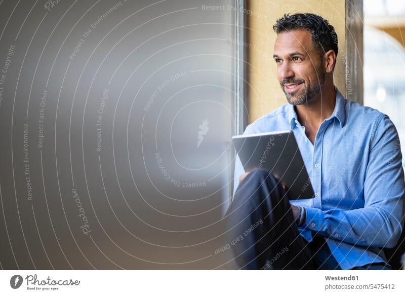 Smiling businessman sitting at the window using tablet indoors indoor shot Interiors indoor shots interior view mobile working accessibility accessible online