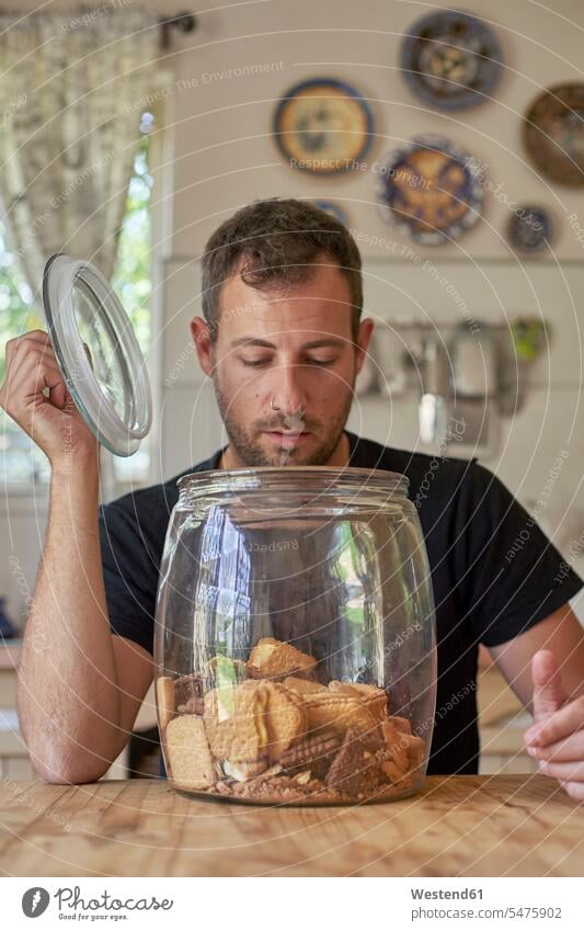 Man sitting in kitchen, looking in cookie jar Tables wood wood table hold Seated at home Alimentation food Food and Drinks Nutrition foods sweet Pastries