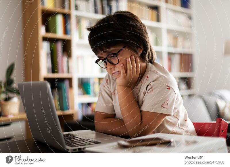 Cute boy with hand on chin looking at laptop while sitting in living room color image colour image indoors indoor shot indoor shots interior interior view