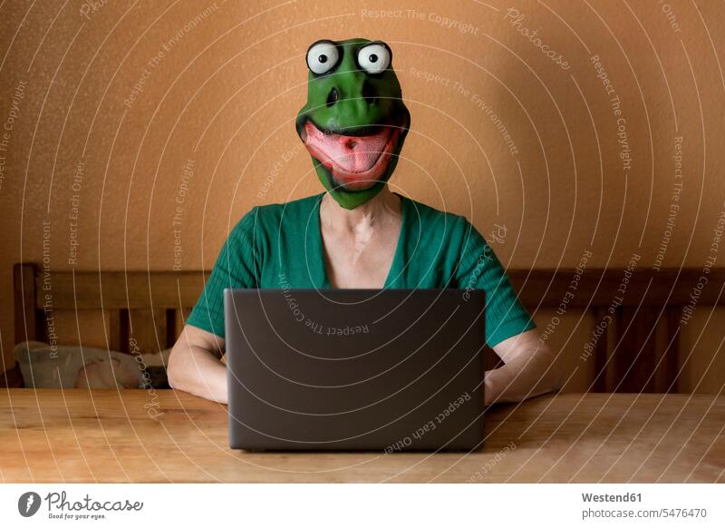 Playful mature woman wearing frog mask while using laptop on table at home during curfew color image colour image Germany indoors indoor shot indoor shots