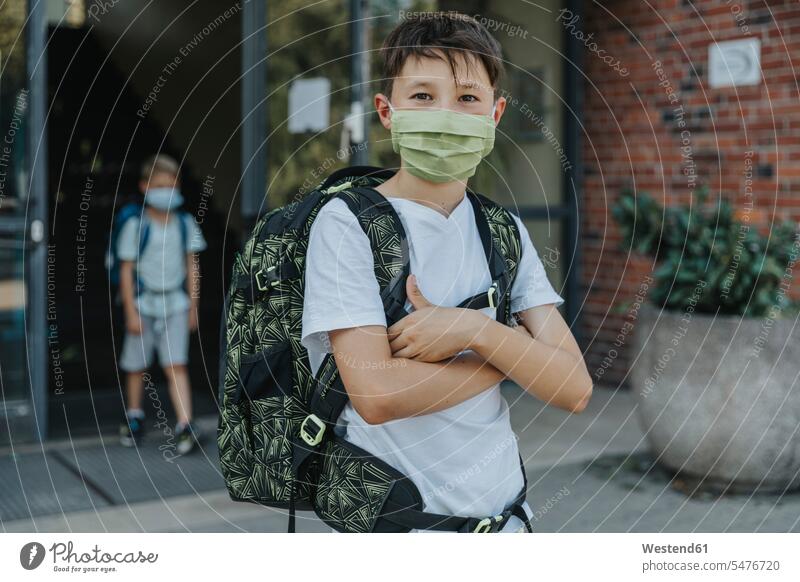 Little boy wearing protective face mask with arms crossed standing in front of school building color image colour image outdoors location shots outdoor shot