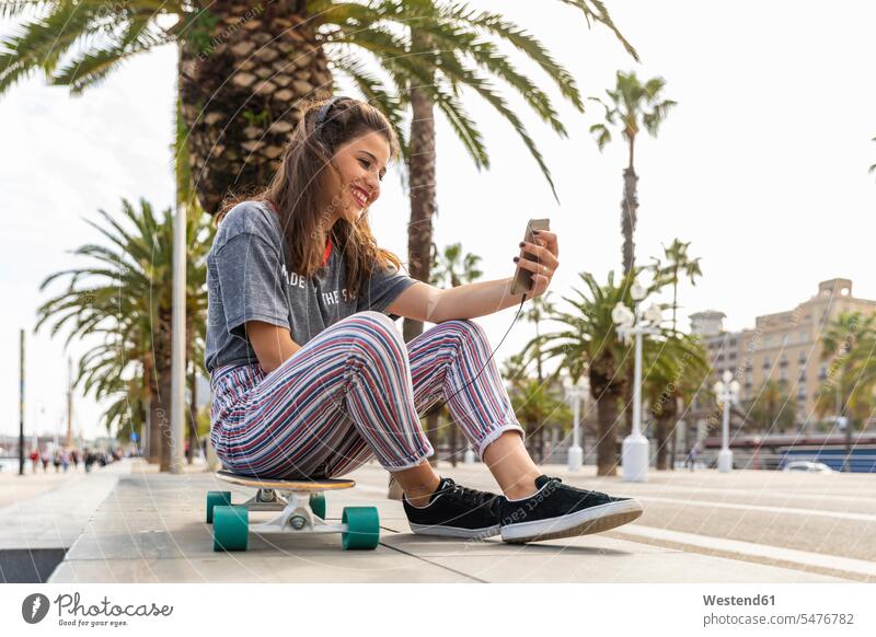 Happy teenage girl sitting on skateboard listening to music with headphones Skate Board skateboards happiness happy hearing headset urban urbanity sky skies