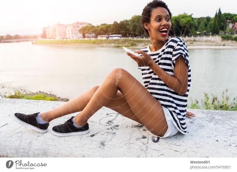 Happy young woman sitting on wall at the riverside holding cell phone females women walls mobile phone mobiles mobile phones Cellphone cell phones Seated