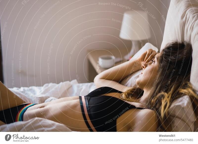 Seductive young brown-haired woman in white bra and panties lying down on  bed at home.. Stock Photo