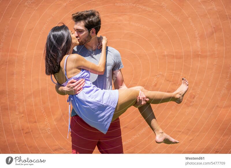 Young man kissing while carrying woman against brown wall at back yard during sunny day color image colour image Spain bonding community leisure activity