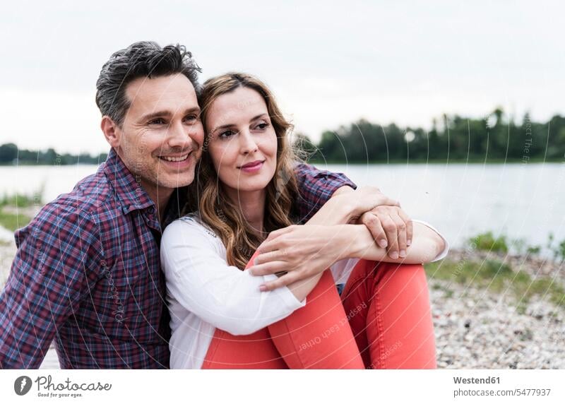 Affectionate happy couple sitting at the riverside Seated riverbank happiness twosomes partnership couples smiling smile River Rivers water's edge waterside