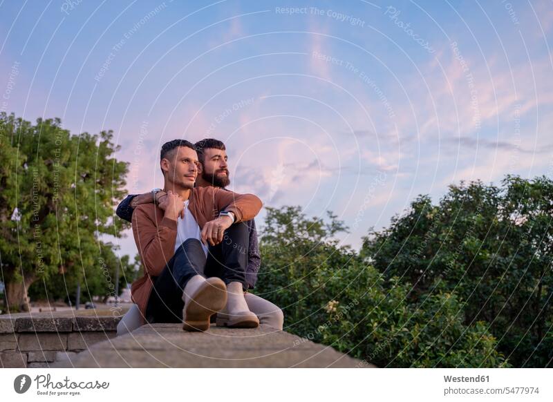 Affectionate gay couple sitting on a wall at sunset associate associates partner partners partnerships touch relax relaxing cuddle snuggle snuggling Seated