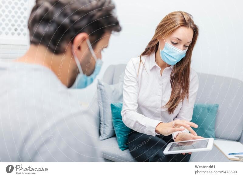 Doctor wearing face mask using digital tablet while sitting by man at office color image colour image indoors indoor shot indoor shots interior interior view