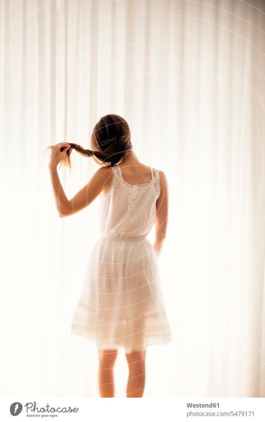 Young woman playing with her hair in front of a white curtain, back view Curtain Curtains Drapery Draperies Selective focus Differential Focus Contour twisting
