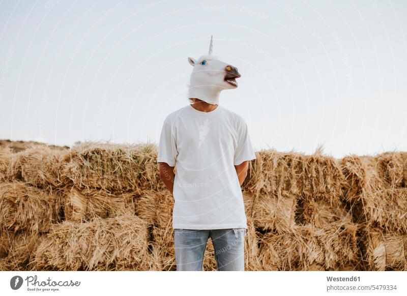 Portrait of young man wearing unicorn mask standing in front of stacked straw outdoors location shots outdoor shot outdoor shots day daylight shot
