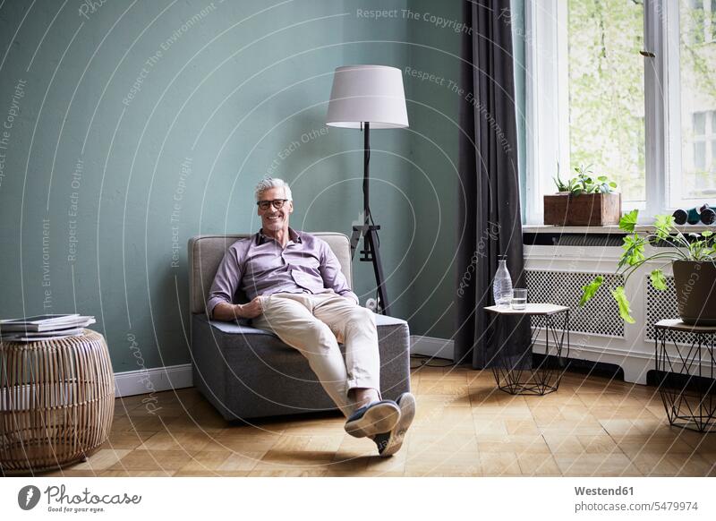 Portrait of smiling mature man sitting on armchair at home Seated men males smile portrait portraits Adults grown-ups grownups adult people persons human being