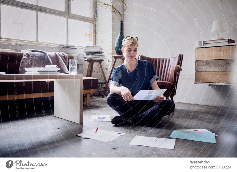 Smiling woman sitting on the floor in a loft looking at papers females women freelancer freelancing lofts Adults grown-ups grownups adult people persons