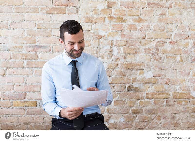 Smiling businessman reading document at brick wall Businessman Business man Businessmen Business men paper documents papers brick walls smiling smile