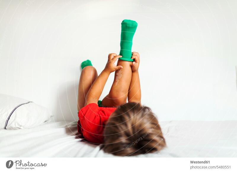 Back view of little boy lying on bed putting on his green socks beds boys males child children kid kids people persons human being humans human beings stocking