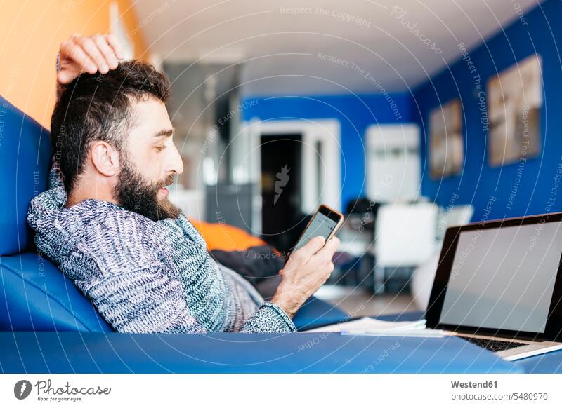 Bearded young man working at home relaxed sitting on the couch, using laptop and mobile content pleased self-employment self-employed freelancer freelancing