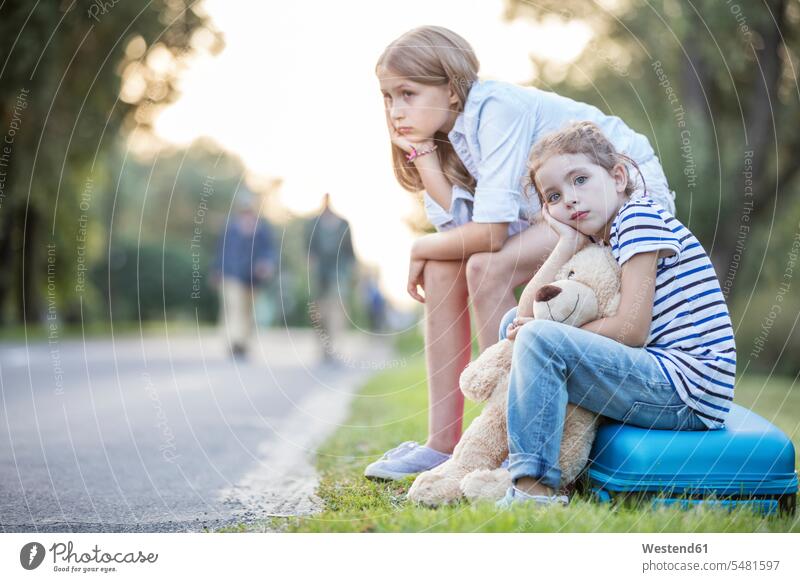 Two girls sitting on suitcase at the roadside waiting suitcases Seated sister sisters Road Side siblings brother and sister brothers and sisters family families