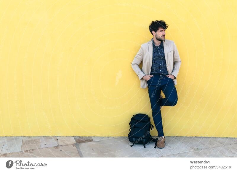 Young man with backpack standing in front of yellow wall men males Adults grown-ups grownups adult people persons human being humans human beings leaning walls