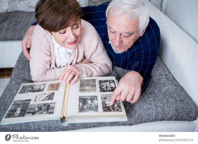 Senior couple lying on couch, looking at photo album photograph photographs photos eyeing senior adults seniors old photograph album photo albums watching