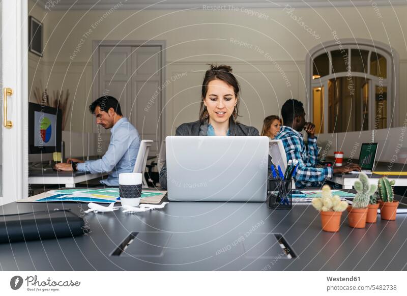 Young businesswoman using laptop, colleagues working in background team sitting Seated At Work Laptop Computers laptops notebook businesswomen business woman
