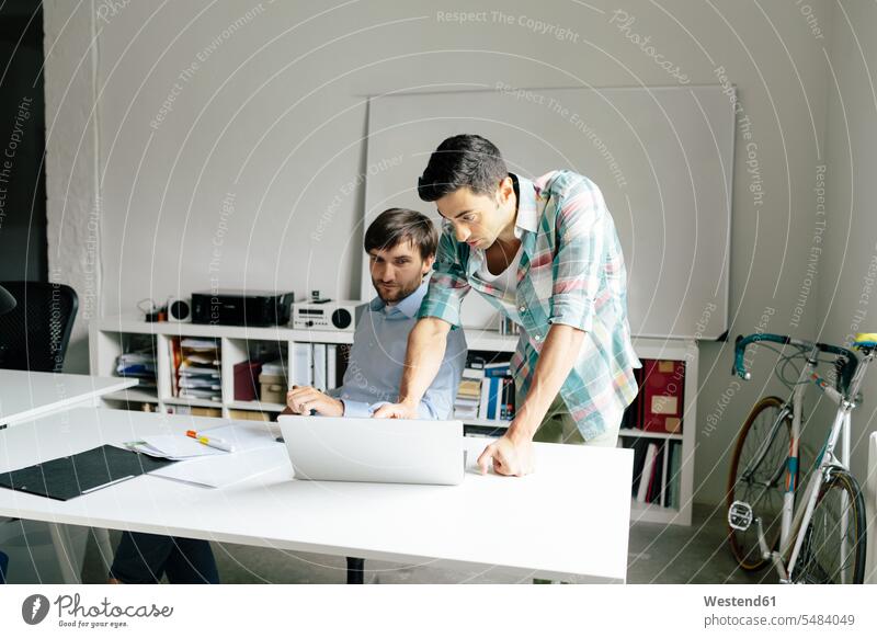 Two businessmen with laptop at desk in office discussing Laptop Computers laptops notebook colleagues offices office room office rooms Businessman Business man