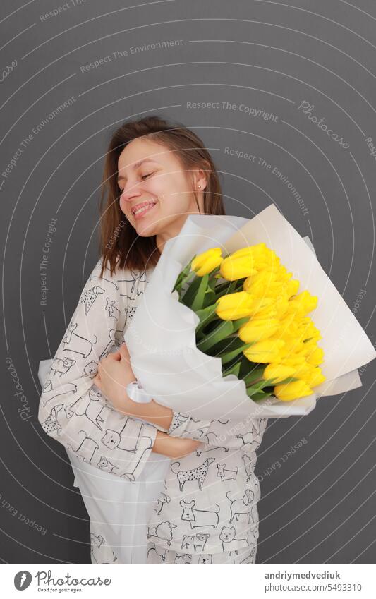 Very nice young happy woman holding big and beautiful blossoming bouquet of fresh yellow tulips flowers on the grey wall background. International Women's Day