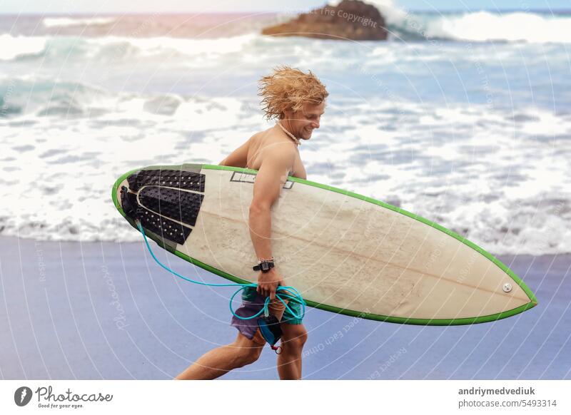 Fit young surfer man with curly blond hair with surfboard goes by the ocean having fun doing extreme water sports, surfing. Travel and healthy lifestyle concept. Sports travel destination.