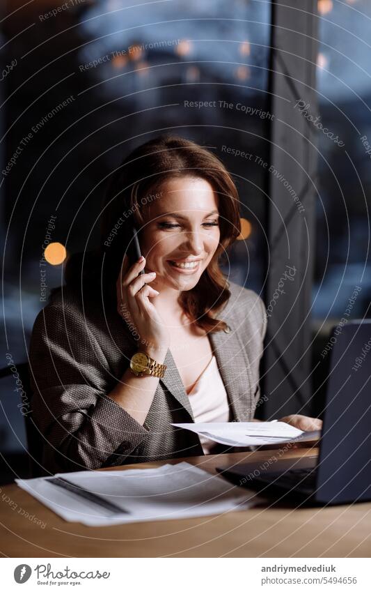 smiling successful beautiful young businesswoman working in cafe. female is talking cellphone while working remotely in elegant wooden interior with many greens. laptop with documents on table.