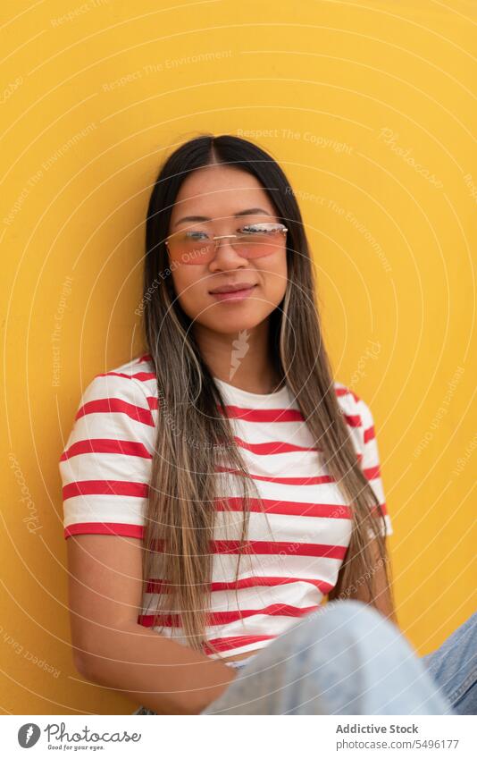Smiling woman in eyeglasses sitting against yellow wall positive happy casual smile eyewear stripe glad optimist fashion pleasure content asian pleasant