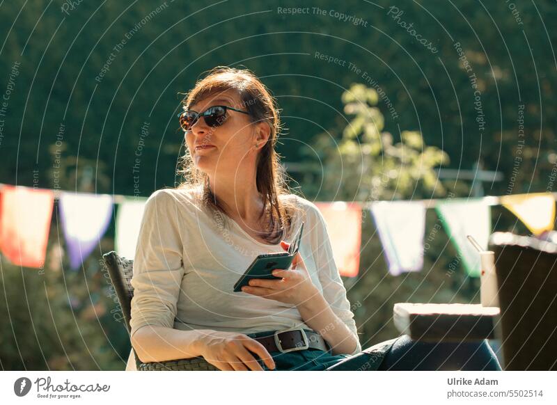 Drinkje bej Inkje |Sunbeams - woman with sunglasses relaxes and enjoys the sunny weather in the beautiful garden. In her hand she holds a smartphone long hairs