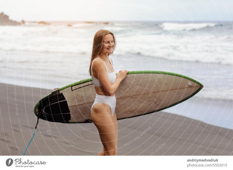 Young surfer woman in bikini going surfing stands with surfboard on the black sandy beach. Girl walks with longboard. Extreme water sport by the ocean, sea. Healthy Active Lifestyle. Summer Vacation
