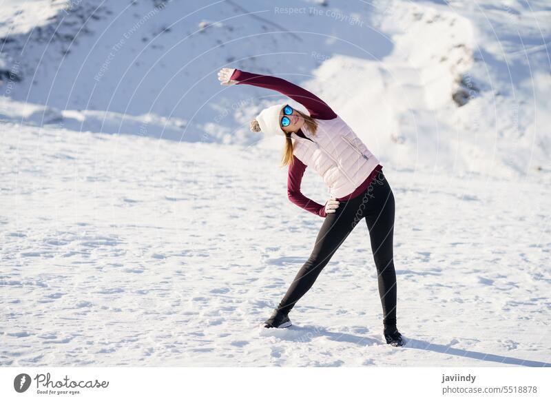 Young woman standing and doing exercise on snow in highlands recreation winter mountain cap stretch landscape female young sunglasses outfit arm raised gesture