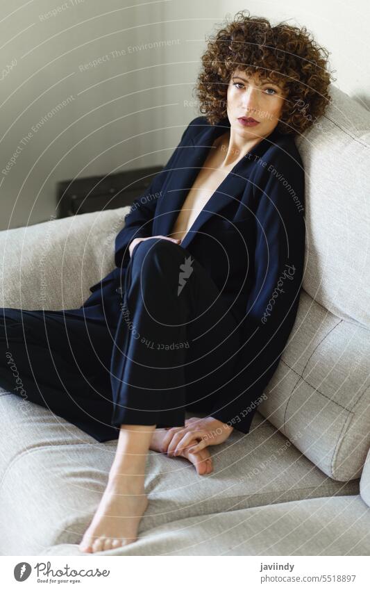 Stylish curly haired woman sitting on sofa wearing black suit confident charismatic posture charm outfit modern female emotionless unemotional adult dark hair