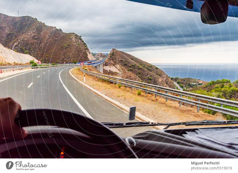 View from the driving position of a truck of a highway that crosses between mountains with the sea in the background. interior inside dashboard hand road cloudy