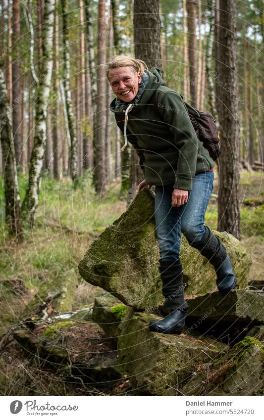 Laughing woman with blond hair, green jacket, blue jeans, gauntlets, rubber boots and brown backpack climbs on granite quarry stones in the middle of mixed forest with birch and conifer trees.