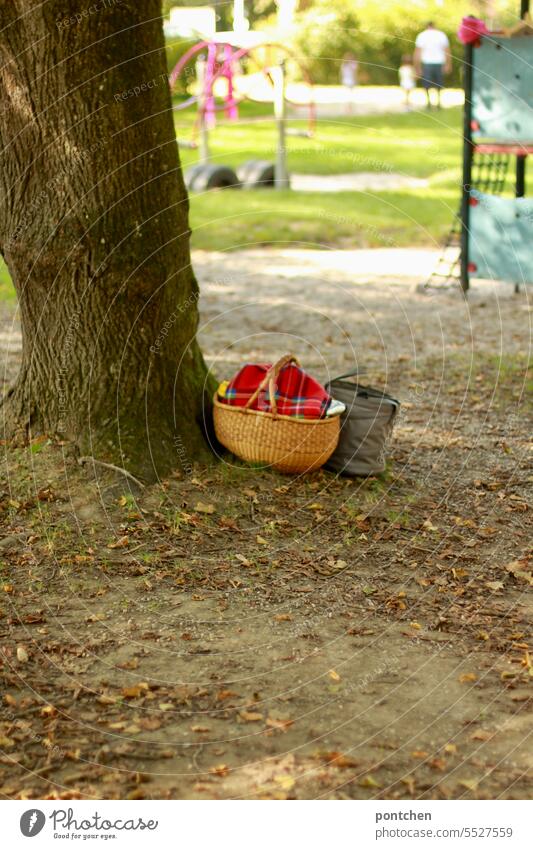 a picnic basket with a checkered blanket stands by a tree in a playground . a father with his two children in the background Playground Supplies Picnic basket