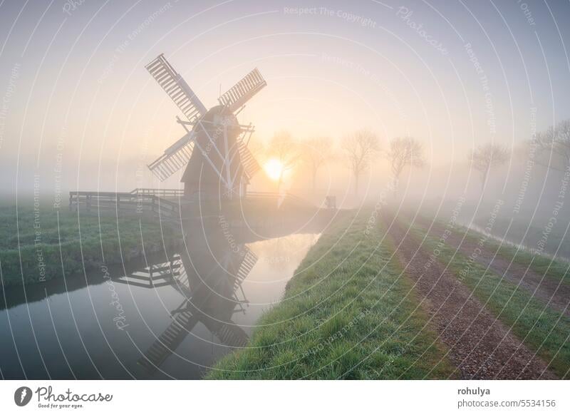 beautiful misty sunrise in Dutch countryside with windmill architecture fog sunlight sunshine river canal water road way path reflection symmetry view scenic