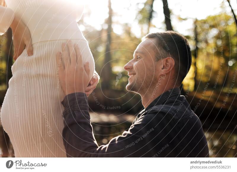 Cropped shot of happy young husband enjoying strokes, talks and kisses tummy of wife, waiting for birth baby outdoors in sunbeams in the city park. Maternity prenatal care and woman pregnancy concept.