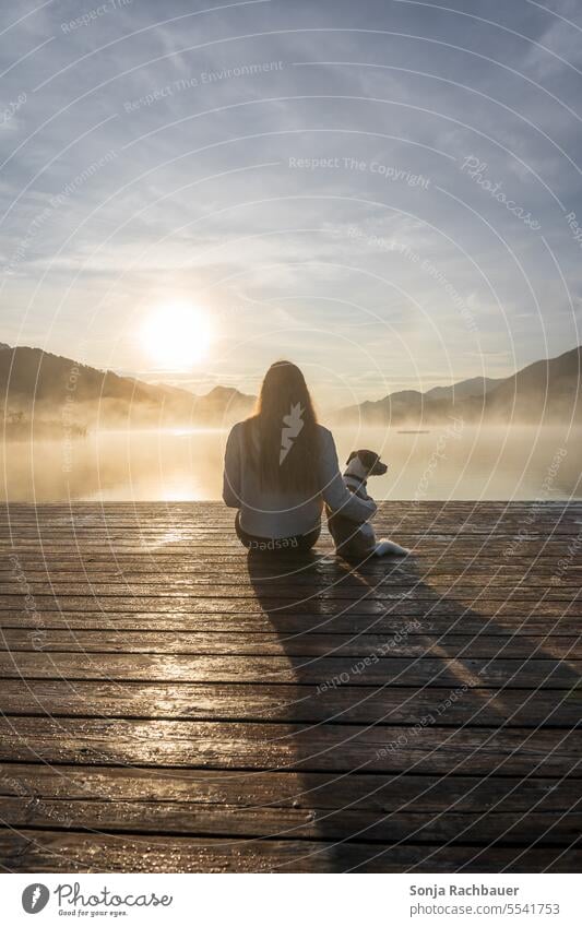 A young woman sits with a small dog on a wooden jetty by a lake. Sunrise, back view. Woman youthful Dog Sit wooden walkway Rear view Fog Autumn Pet Lifestyle